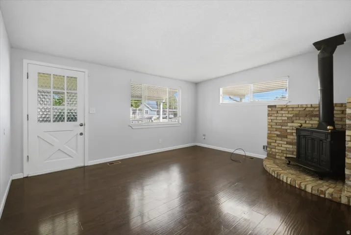 Wood-finish flooring, brick hearth, black wood-burning stove, white textured-glass door, and multiple windows with horizontal blinds