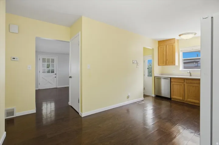 Dark wood-finish flooring throughout, light-toned walls, and a kitchen area featuring wood cabinetry, a stainless steel dishwasher, and a window