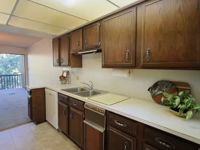 Wood cabinetry with chrome hardware, dual basin stainless steel sink, white countertops, built-in dishwasher, and tile flooring