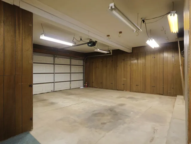 Concrete flooring, wood-paneled walls, built-in storage cabinets, overhead fluorescent lighting, and an automatic garage door