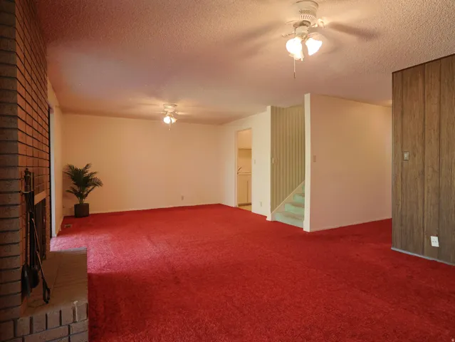 Exposed brick fireplace with hearth, two ceiling fans with light kits, staircase with carpeted treads, and wood-paneled wall accent