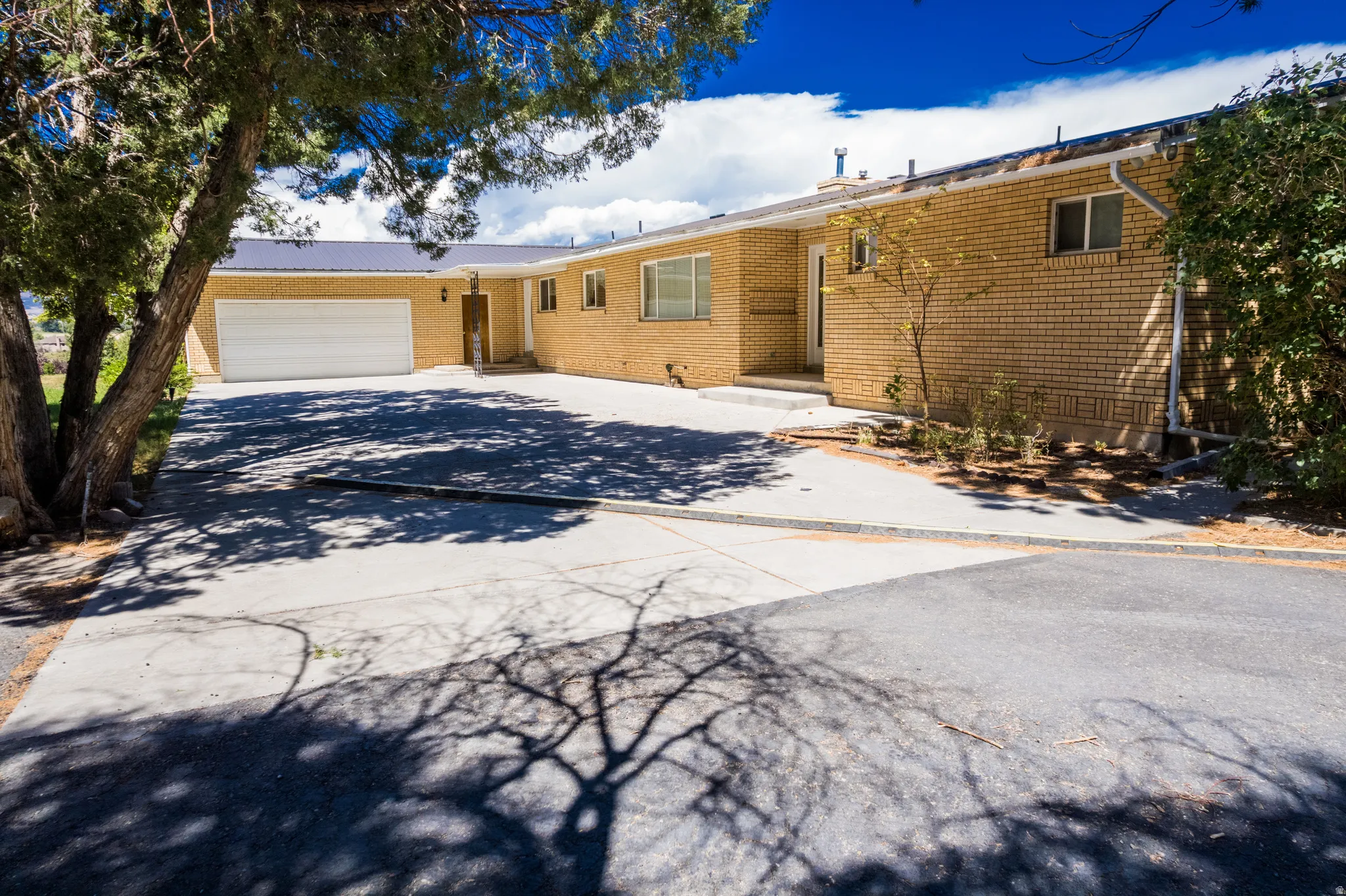 Brick exterior with a metal roof, an attached garage, a paved driveway, and mature landscaping