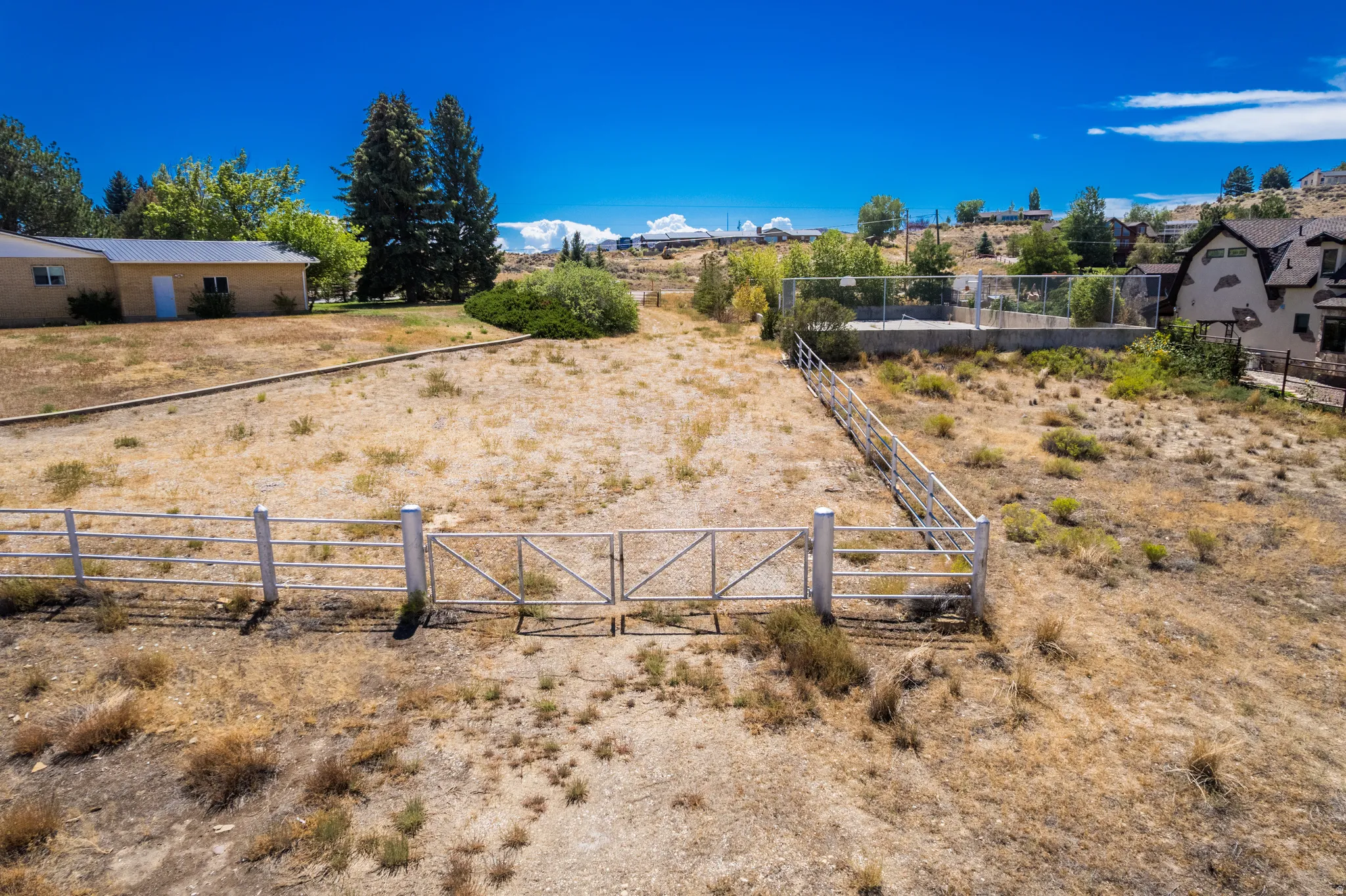 Expansive lot featuring a perimeter fence with a double gate, mature trees, and a distant building with a light-colored exterior