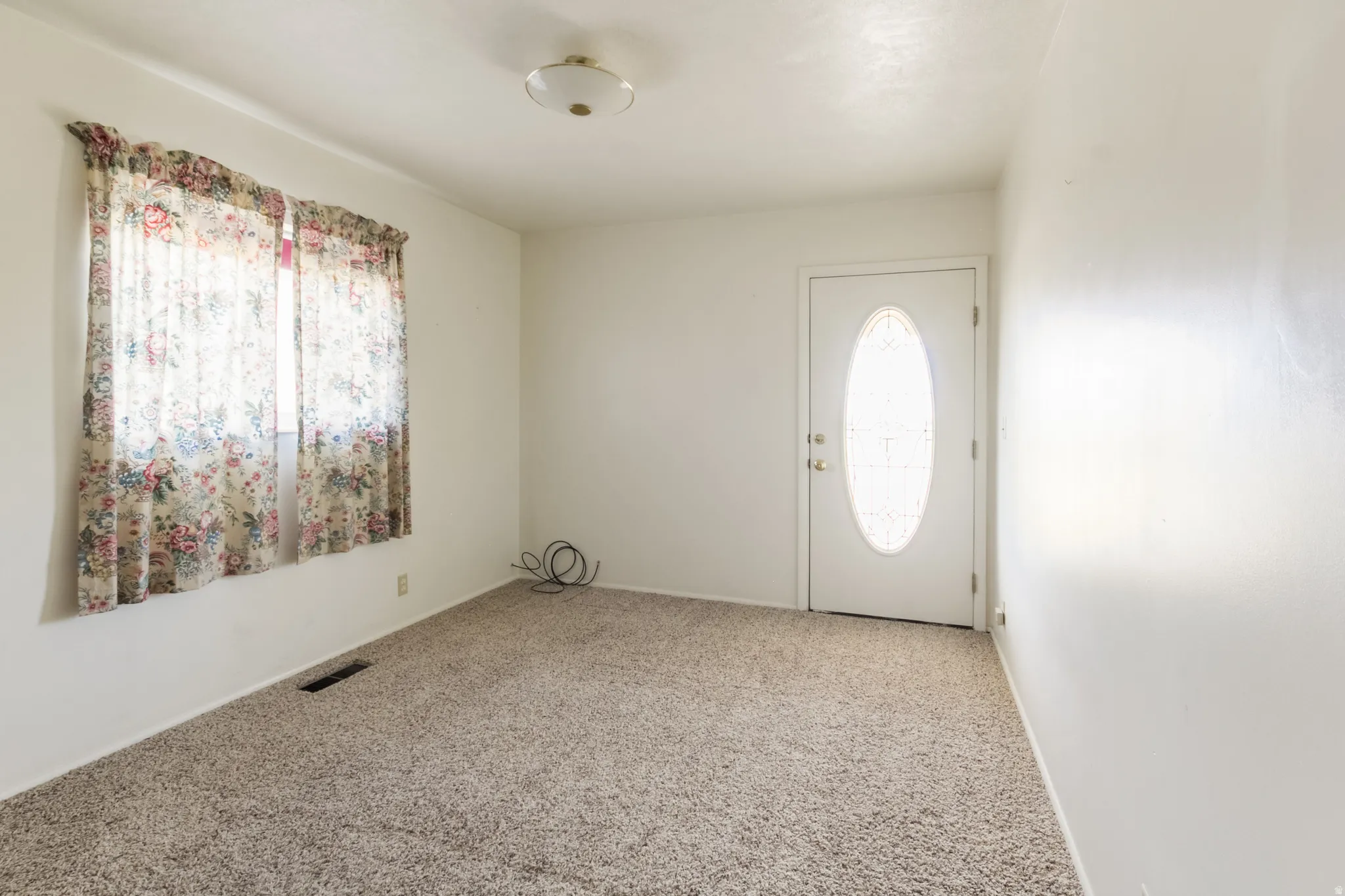 White interior walls, neutral carpet flooring, ceiling-mounted light fixture, and an entry door with an oval-shaped glass insert