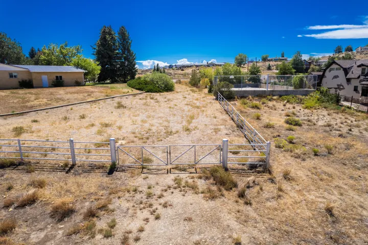 Expansive lot featuring a perimeter fence with a double gate, mature trees, and a distant building with a light-colored exterior