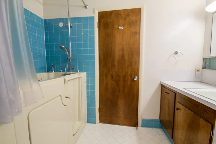 Walk-in tub with chrome fixtures, blue tiled shower surround, wood-finish vanity with white countertop, and tiled flooring
