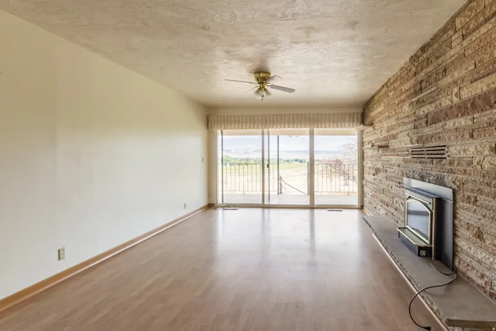 Wood-finish flooring, full-height stone accent wall, built-in fireplace, sliding glass doors, and a ceiling fan