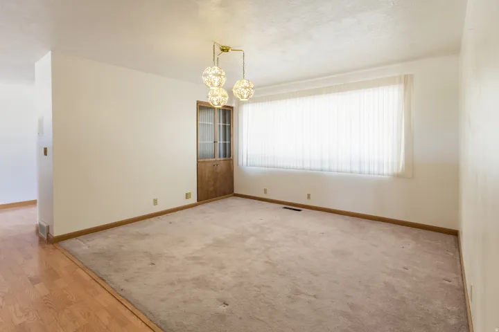 Spacious room featuring a wide window with vertical blinds, a decorative ceiling light fixture, a wood-frame door with frosted glass, and wood-finish flooring in an adjacent area