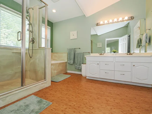 Spacious bathroom featuring a glass-enclosed shower, built-in tub with stone-look surround, a dual vanity with ample storage, a large wall-mounted mirror, and wood-finish flooring