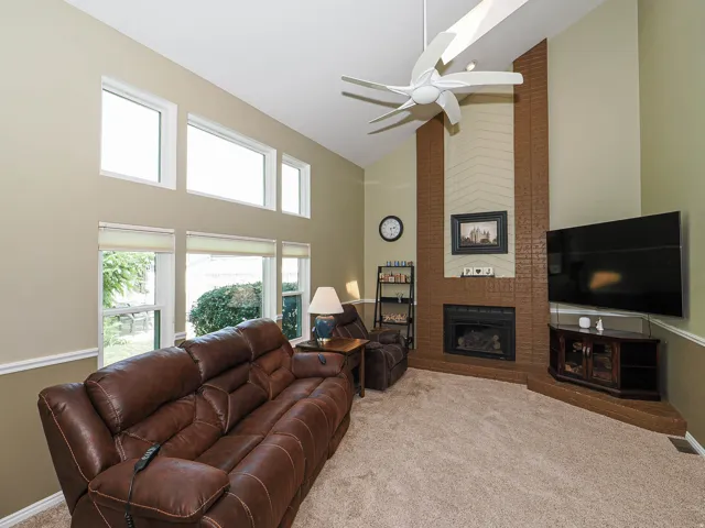Vaulted ceiling with a contemporary ceiling fan, brick fireplace surround with mantel, multiple transom windows, and neutral carpeting