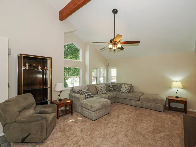 Vaulted ceiling with exposed wood beam, ceiling fan with light fixture, multiple windows, and plush carpeting