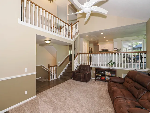 Vaulted ceiling with a ceiling fan, an upper-level balcony with white balusters and wood-finish railing, a kitchen area with white cabinetry, and wood-finish flooring in the entryway