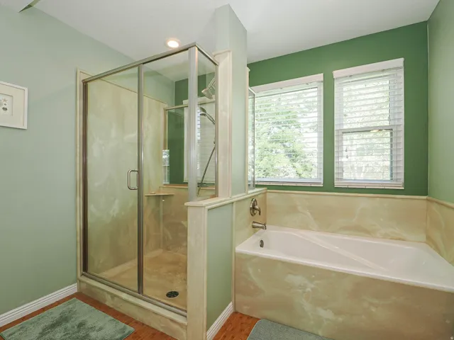 Glass-enclosed shower with a built-in bench, separate soaking tub with stone-finish surround, and two windows with blinds