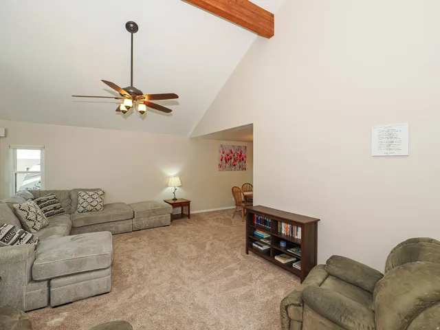 Vaulted ceiling with exposed wood beam, ceiling fan with integrated lighting, window with white trim, and neutral-toned carpet