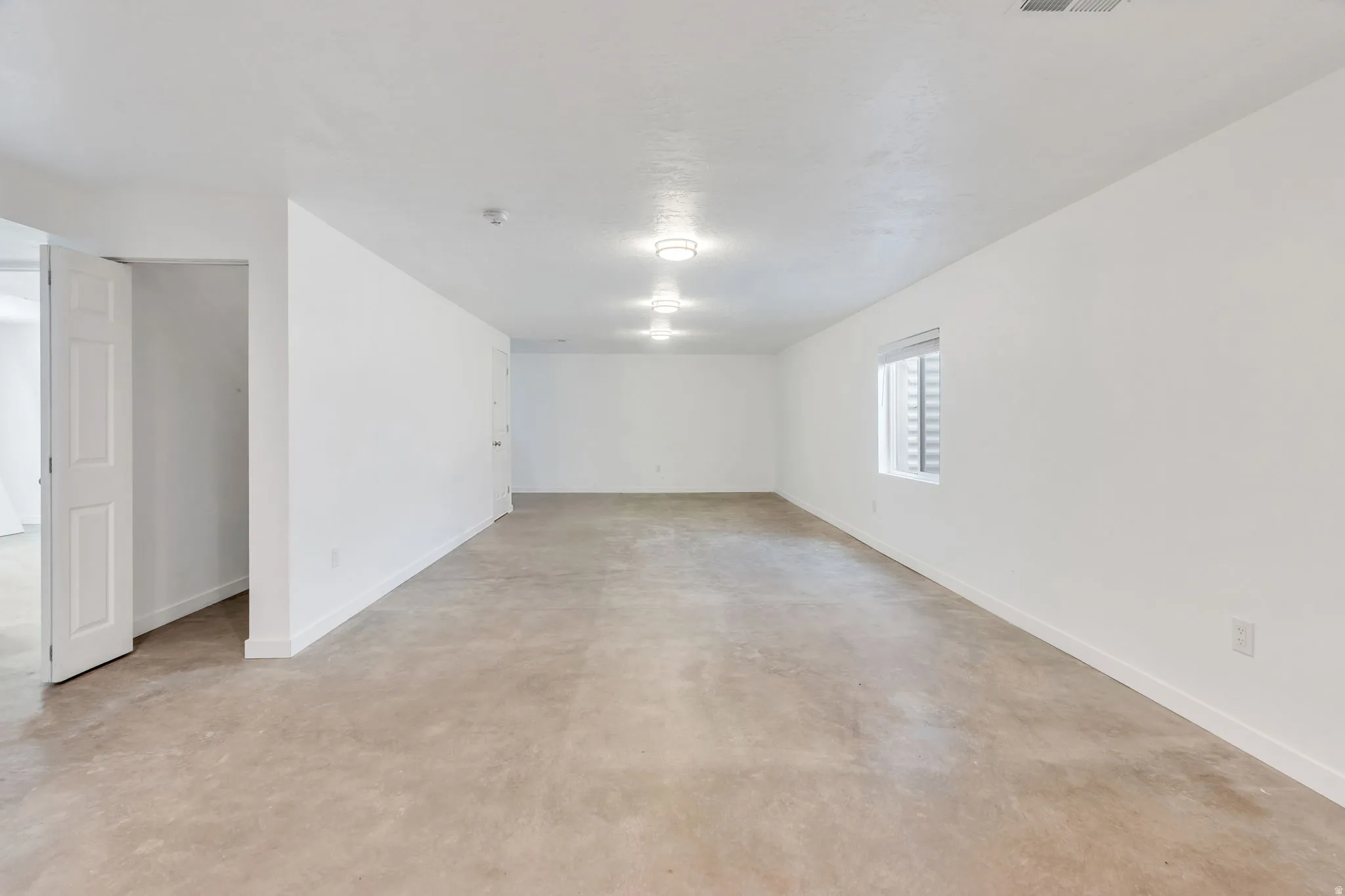 Expansive open space with light-colored carpeting, multiple overhead light fixtures, a window with horizontal blinds, and white painted walls