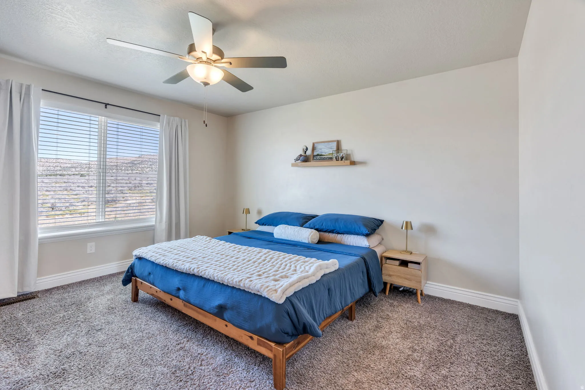 Neutral wall paint, textured carpeting, window with blinds, ceiling fan with integrated lighting, and a wood-finish floating shelf