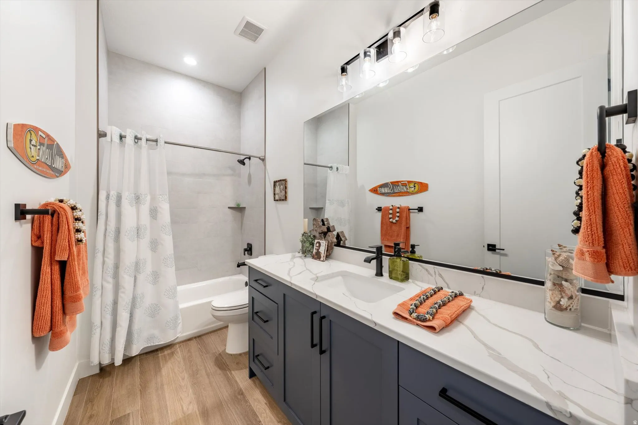 Wood-finish flooring, a white bathtub with light-colored tile surround, a dark vanity with a white countertop, and a large wall-mounted mirror