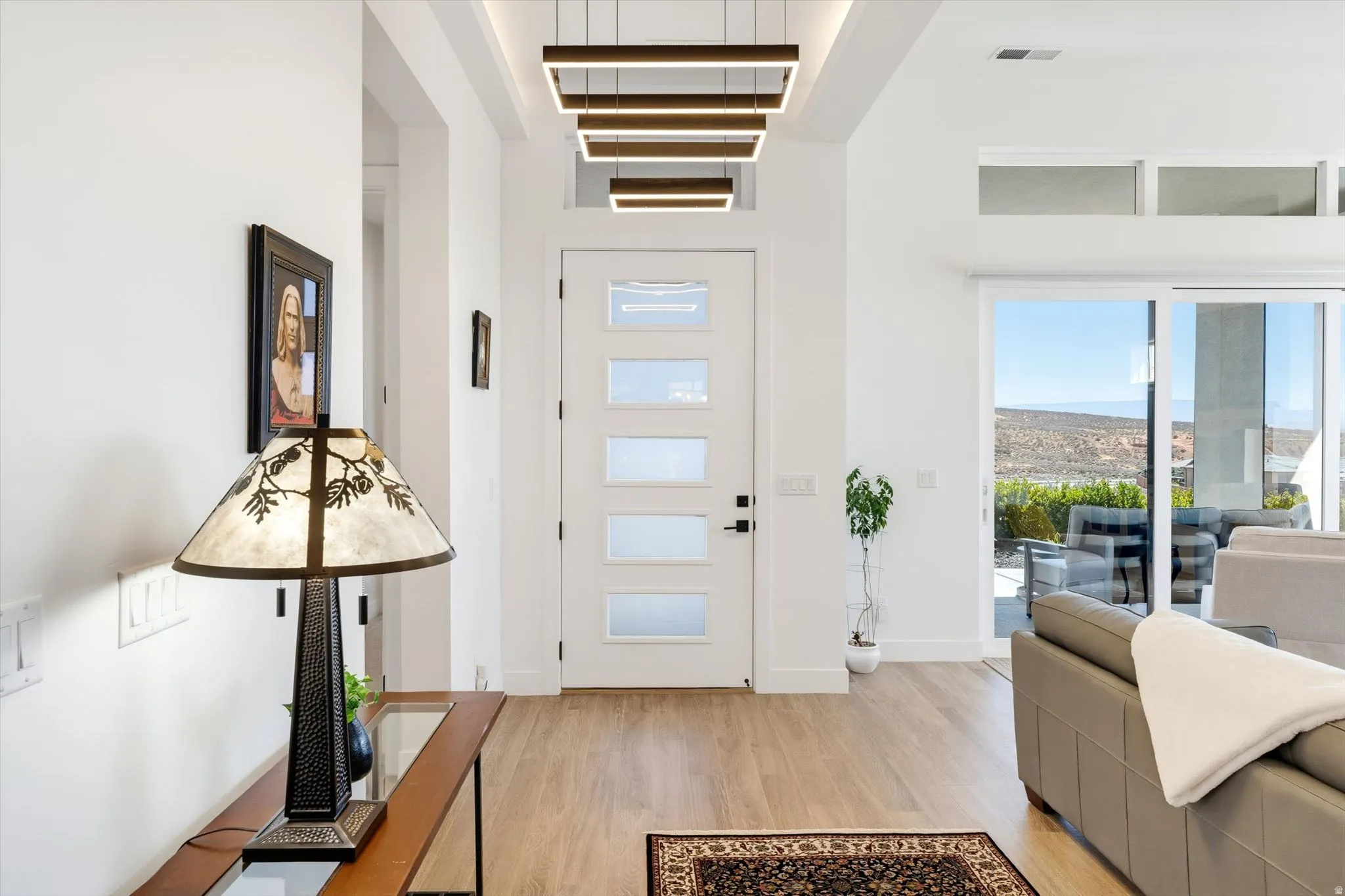 Modern entry door with frosted panels, contemporary linear pendant lighting, wood-finish flooring, and a sliding glass door with transom windows