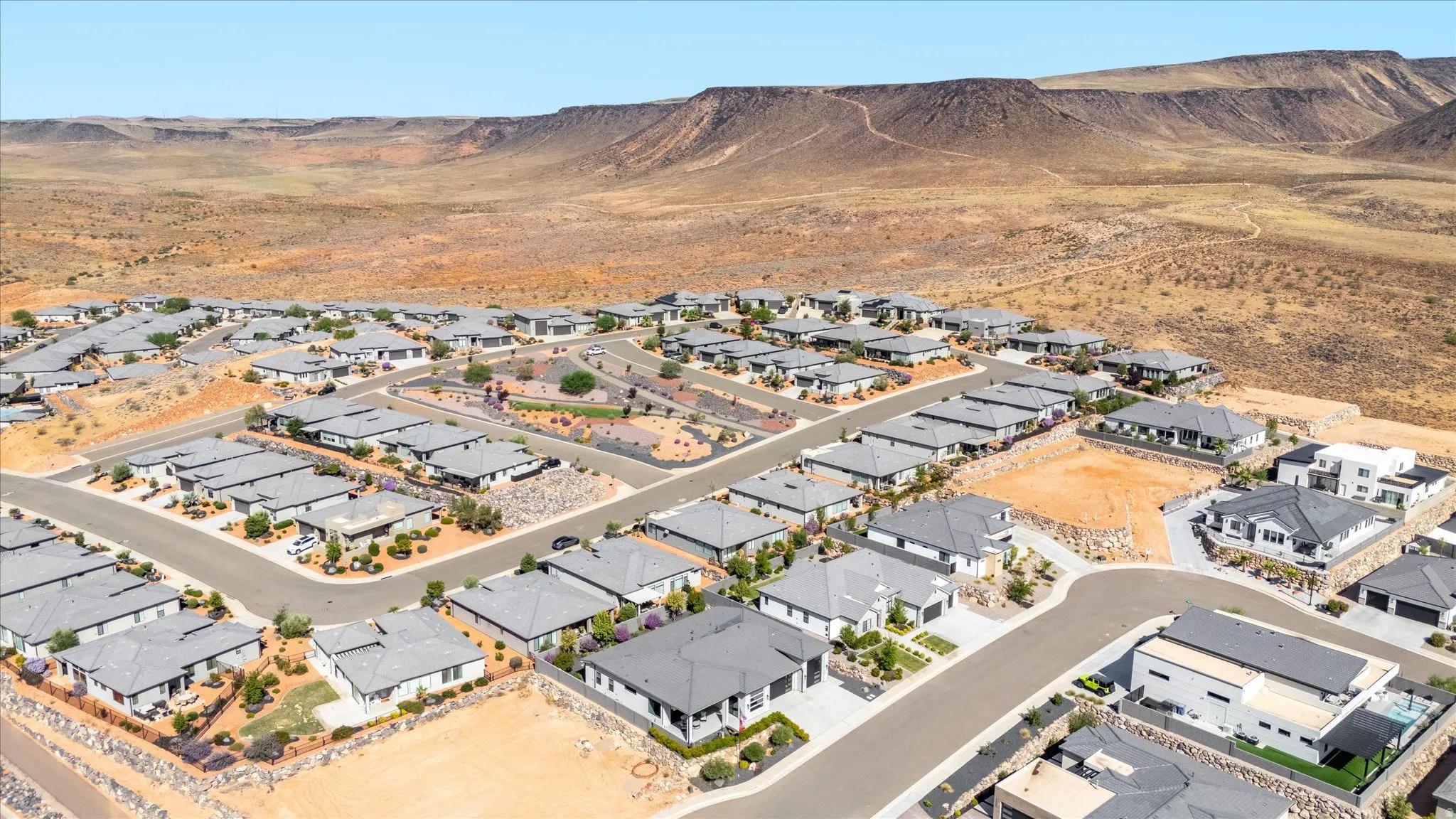 Community of single-story residences with grey tile roofs and arid landscaping, featuring paved streets and desert terrain backdrop