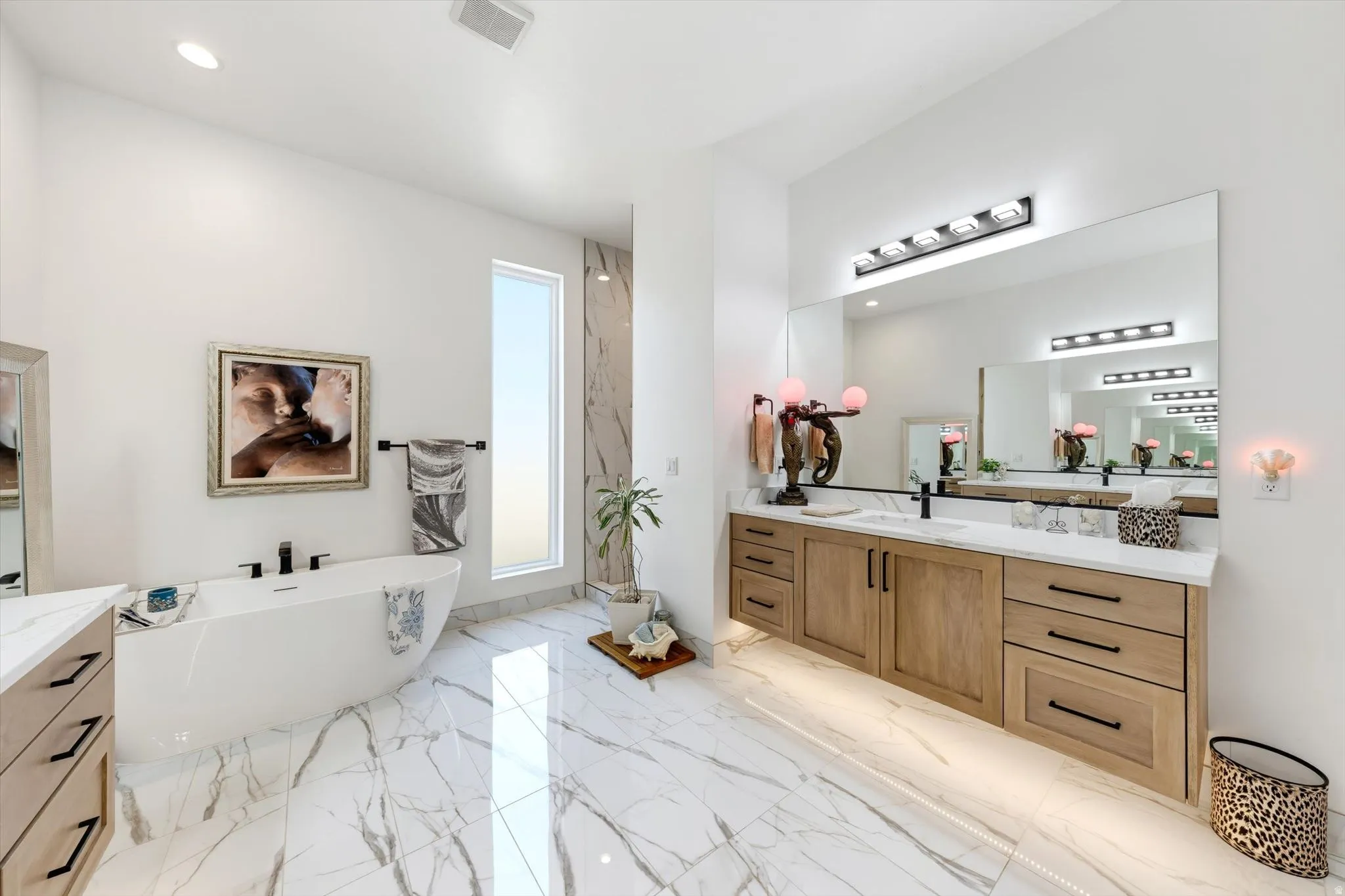 Freestanding soaking tub, dual floating wood-finish vanities with undermount lighting, integrated large mirror, and porcelain tile flooring and shower surround