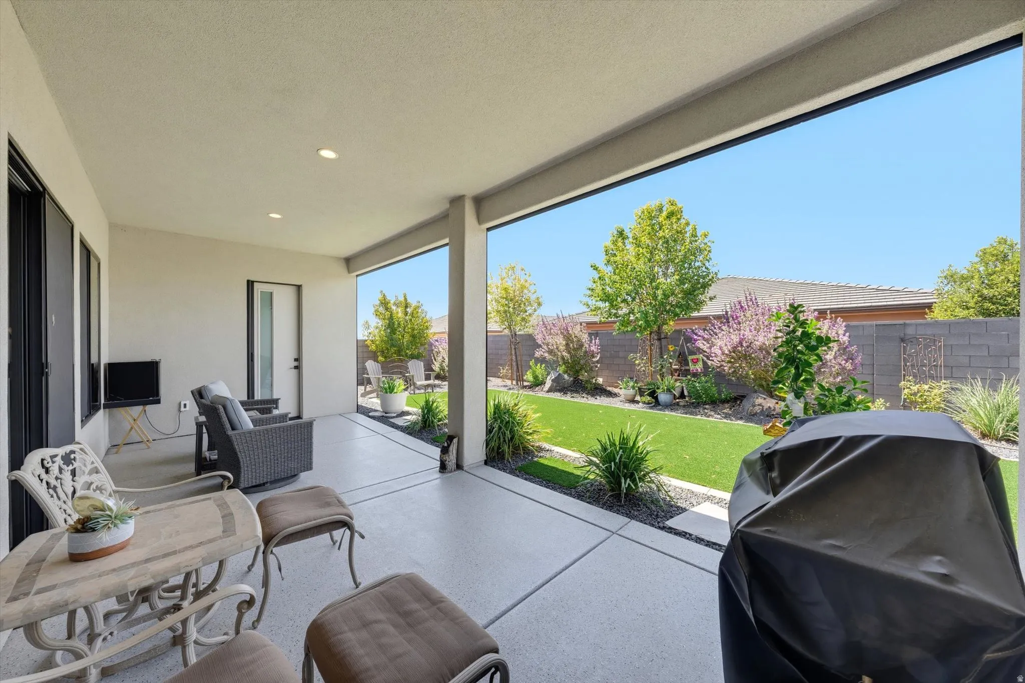 Covered patio with recessed lighting, concrete flooring, and a landscaped backyard with artificial turf and mature plantings