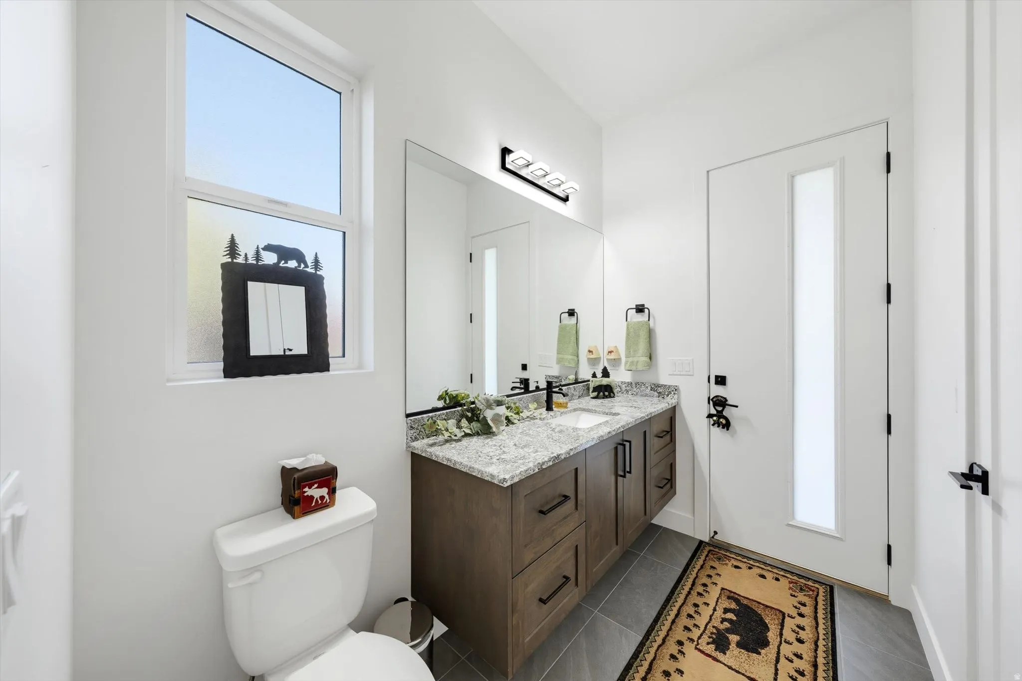 White interior walls with a window, a vanity with a light-colored countertop, a built-in mirror, and dark-finish cabinetry with black hardware