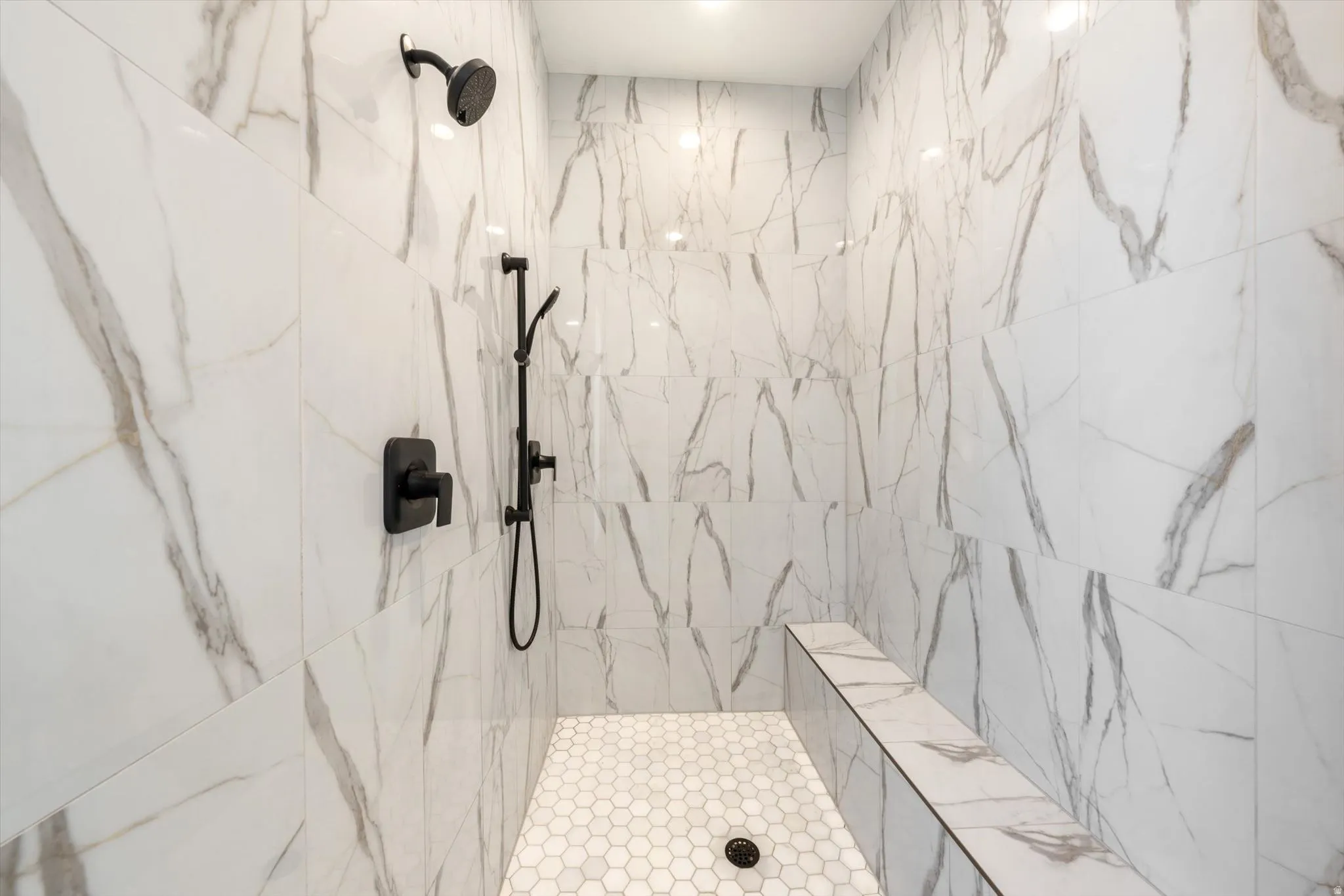 Shower enclosure with marble-look wall tiling, hexagonal floor tiling, matte black fixtures, and a built-in bench