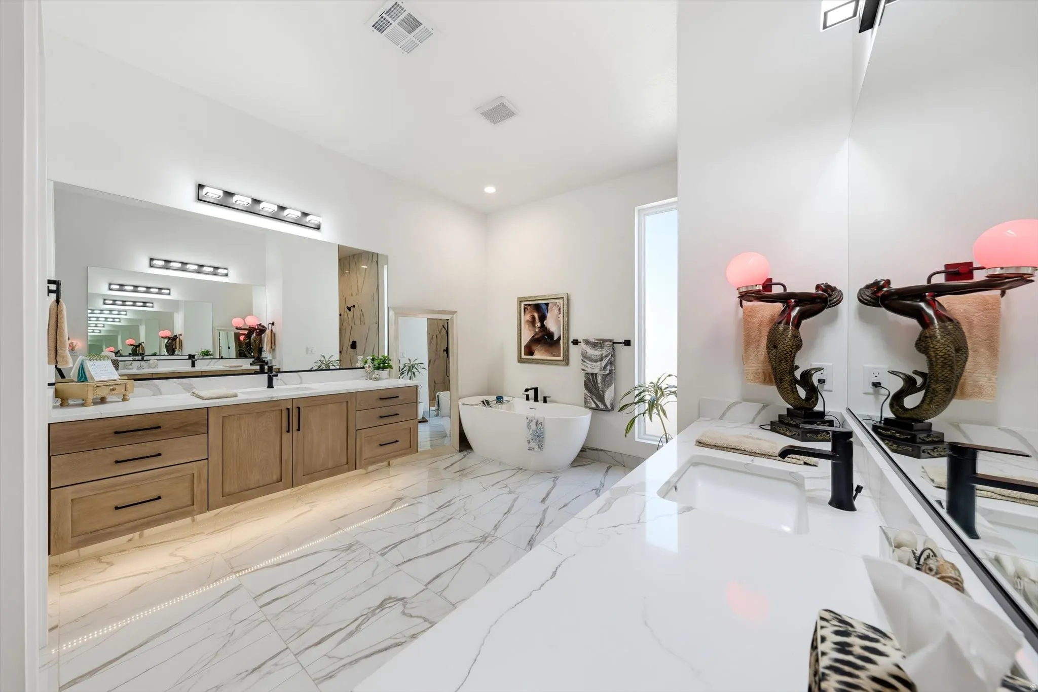Spacious bathroom featuring white marble-look flooring, dual vanities with light wood-finish cabinetry, a freestanding soaking tub, and a window