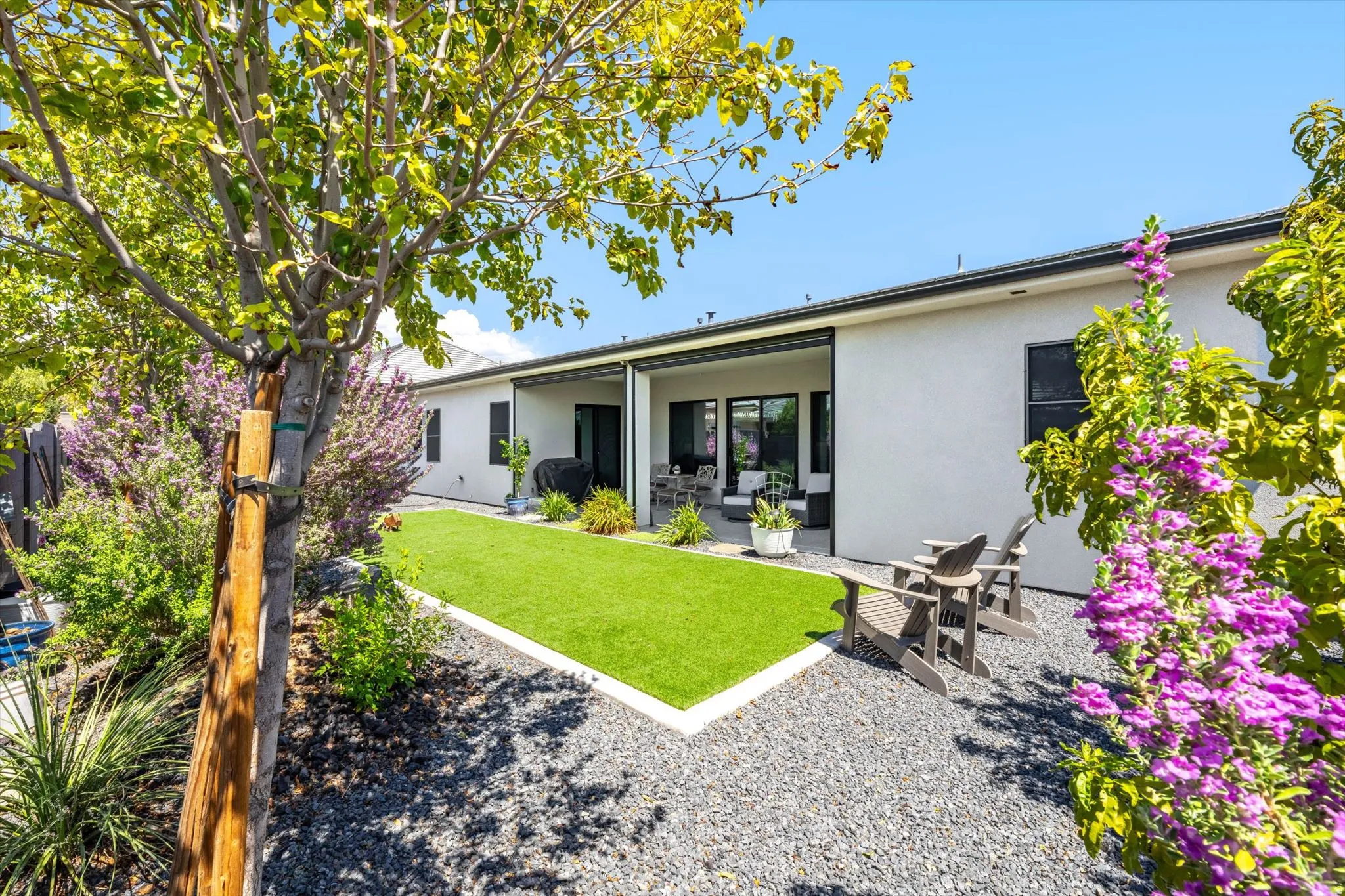 Expansive backyard featuring artificial turf, dark gravel landscaping, mature trees, and a covered patio area with dark-framed sliding glass doors