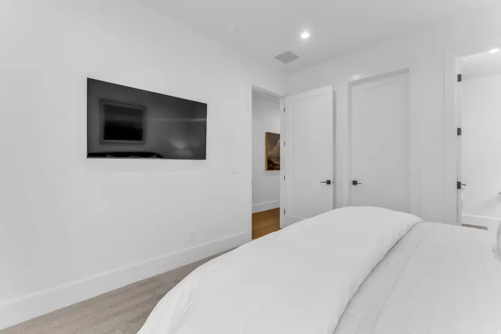 Modern primary bedroom featuring a wall-mounted flat-screen television, wood-finish flooring, recessed ceiling lighting, and multiple closet doors with contemporary hardware