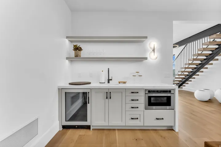 Built-in wet bar with light gray cabinetry, under-counter refrigerator, stainless steel microwave, and floating shelves