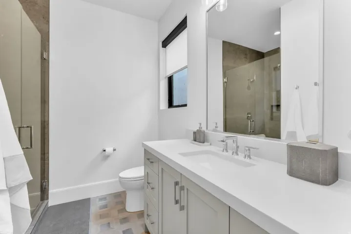 Glass-enclosed shower with a tiled surround, a toilet, a vanity with an integrated sink, a large mirror, and a window with a roller shade