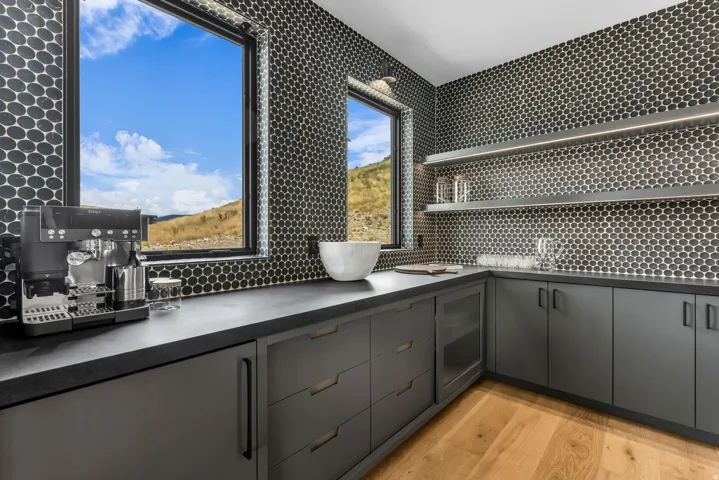 Custom cabinetry with integrated handles, durable countertop surfaces, black penny tile backsplash, two built-in display shelves with lighting, and light wood-finish flooring