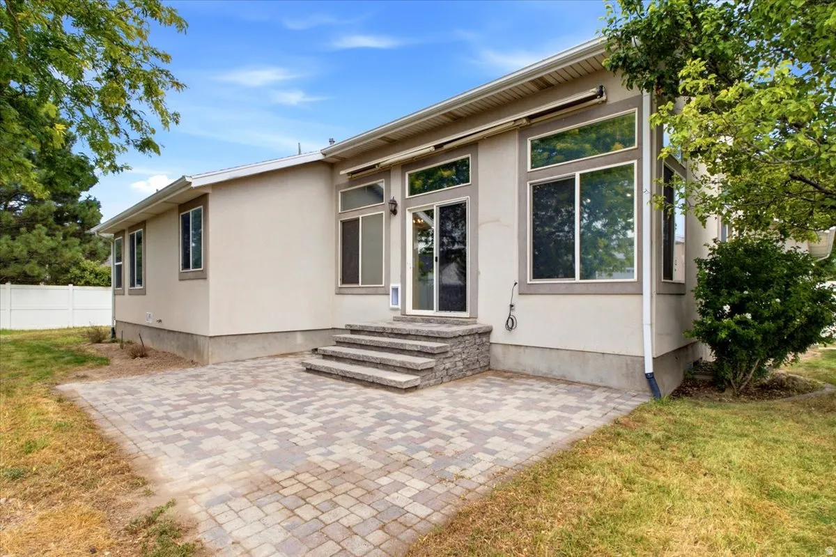 Spacious paver patio with a stacked stone staircase leading to multiple glass-paned doors and large windows, framed by mature trees and a vinyl fence and Open Space Behind.