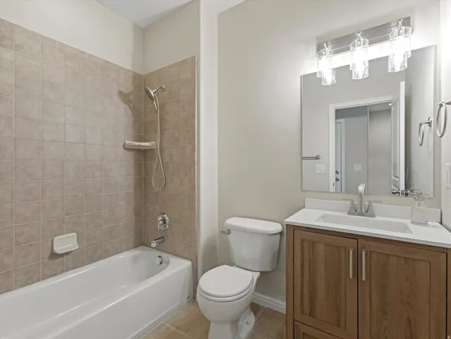 Bathtub with a tiled surround, wood-finish vanity with a white countertop, wall-mounted mirror, and a three-light fixture