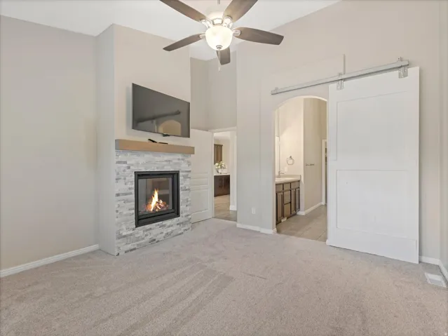 Primary suite, carpeted room featuring a built-in fireplace with a stone surround and wood mantle, vaulted ceiling, ceiling fan, and barn door entry to an arched doorway
