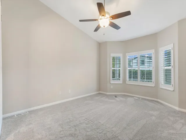 Primary suite, vaulted ceiling, ceiling fan with integrated lighting, three windows with plantation shutters, light gray carpet, and neutral wall paint
