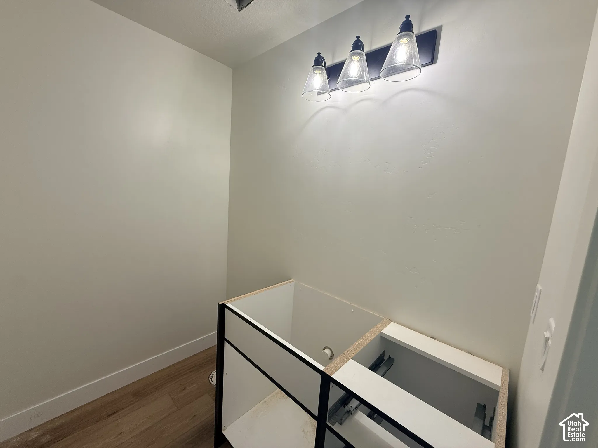 Wall-mounted three-light fixture with clear glass conical shades and matte black hardware, accompanied by a double-basin vanity cabinet featuring a matte black frame and wood-finish flooring