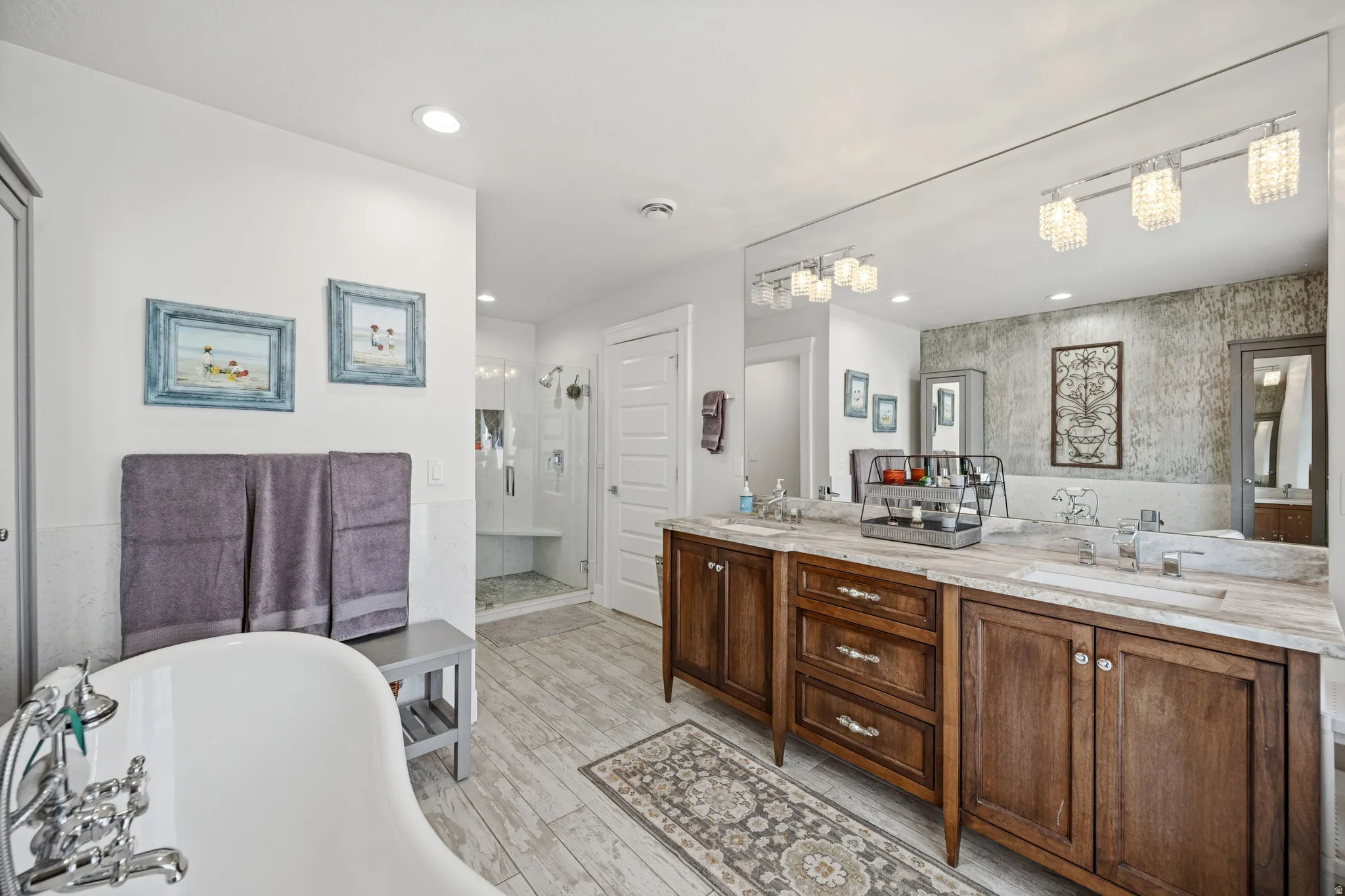 Freestanding bathtub with chrome fixtures, wood-finish flooring, dual vanity with wood cabinetry and undermount sinks, large mirror, and recessed lighting