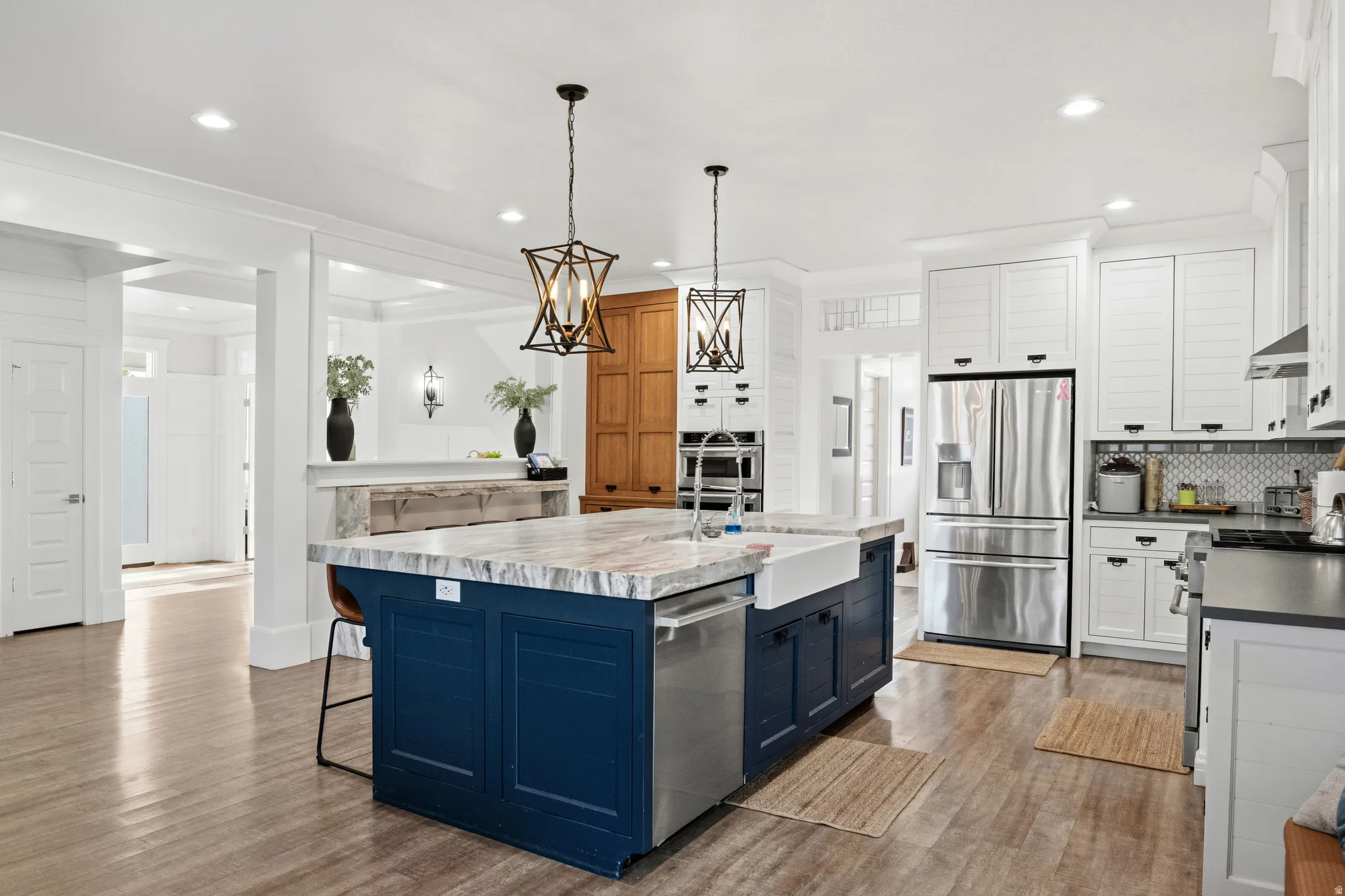 Expansive kitchen featuring a large central island with a farmhouse sink, stainless steel appliances, white perimeter cabinetry, wood-finish flooring, and pendant lighting