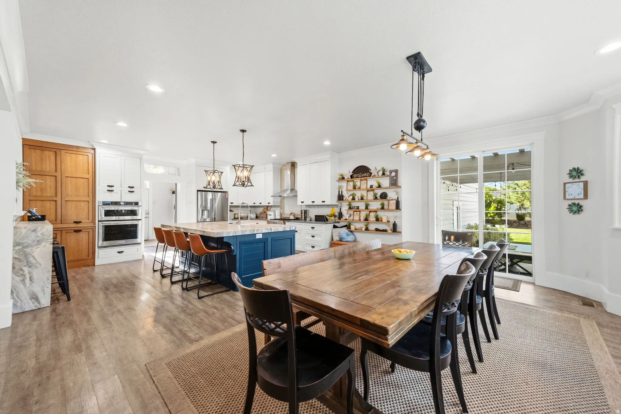 Expansive open-concept kitchen and dining area featuring light wood-finish flooring, a blue kitchen island with seating, stainless steel appliances, and a dining area with direct access to the outdoor space through sliding glass doors