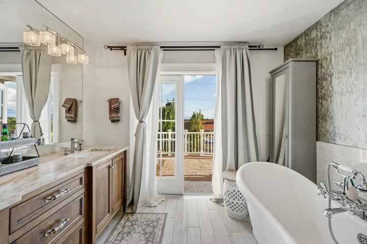 White freestanding tub, wood-finish vanity with dual sinks, stone-finish countertop, and light wood-finish flooring