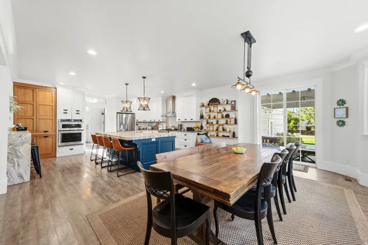 Expansive open-concept kitchen and dining area featuring light wood-finish flooring, a blue kitchen island with seating, stainless steel appliances, and a dining area with direct access to the outdoor space through sliding glass doors