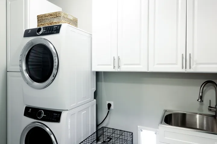 Stacked washer and dryer, white cabinetry with silver hardware, and a stainless steel utility sink with a gooseneck faucet