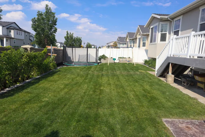 Expansive lawn area with a privacy fence, mature perimeter shrubs, a concrete patio, and exterior stairs with a white railing