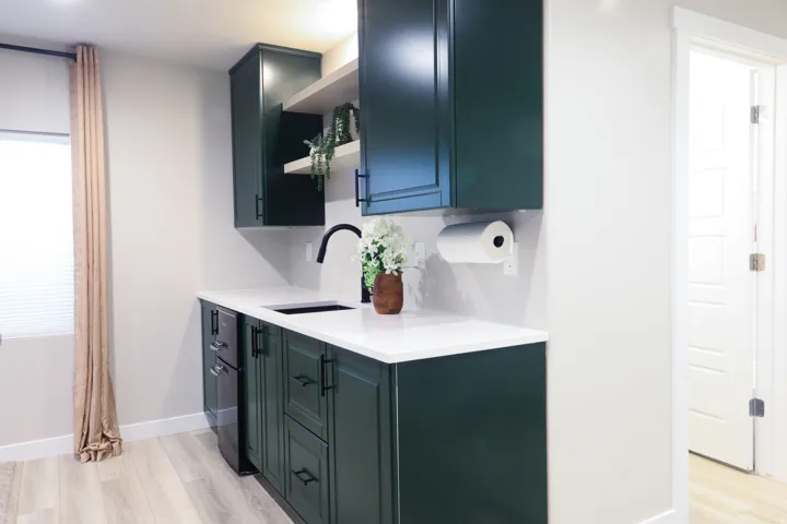 Dark green cabinetry with black hardware, white countertops, built-in sink with matte black faucet, wood-finish flooring, and open shelving