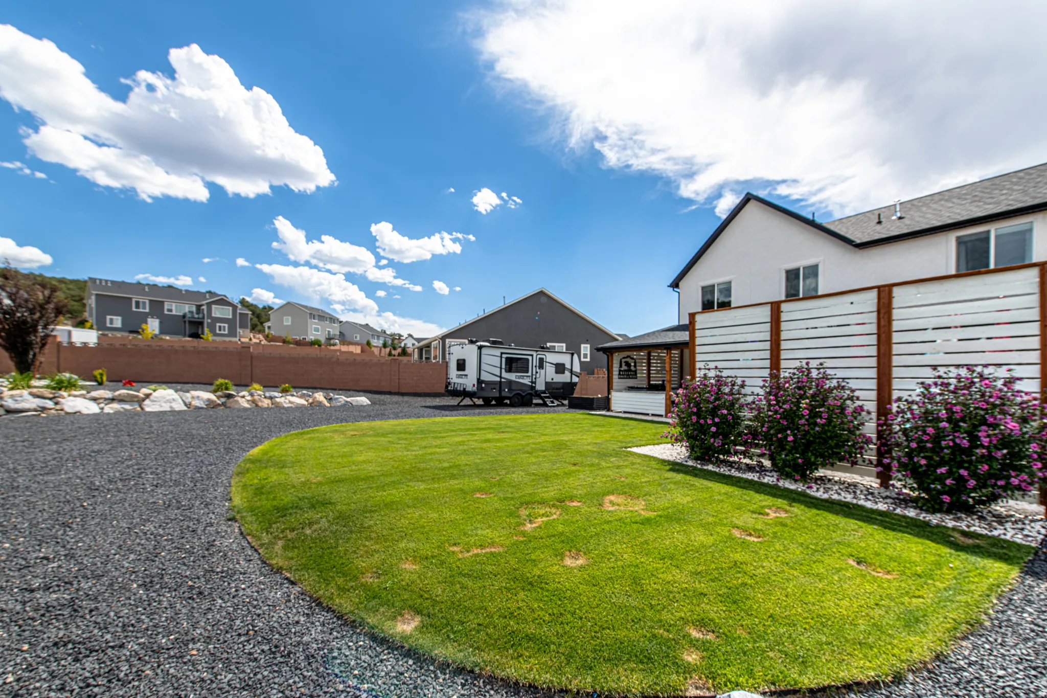 Expansive gravel driveway, manicured lawn, white and wood privacy fence, decorative planted shrubs, and a stucco exterior