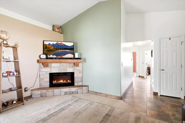 Stone-clad fireplace with a wood mantel, vaulted ceiling, carpeted flooring, and tiled flooring