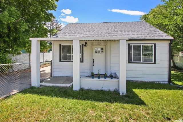 White siding exterior, covered front porch, dark-trimmed windows, gray shingle roof, and a chain-link fence