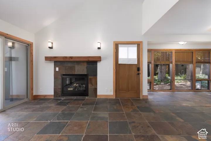 Vaulted ceiling with sconce lighting, natural wood trim, fireplace with a stone surround and wood mantel, large format tile flooring, and abundant natural light from multiple windows